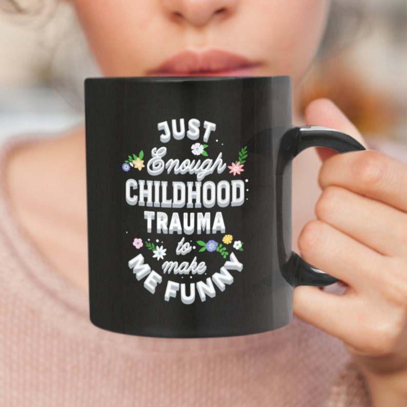 Puede incluir: Taza de caf&eacute; negra con texto blanco que dice "Just Enough Childhood Trauma to Make Me Funny" con un dise&ntilde;o floral alrededor del texto.