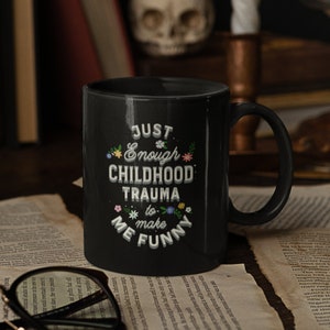 Puede incluir: Taza de caf&eacute; negra con texto blanco que dice "Just Enough Childhood Trauma to Make Me Funny" con peque&ntilde;os detalles florales.