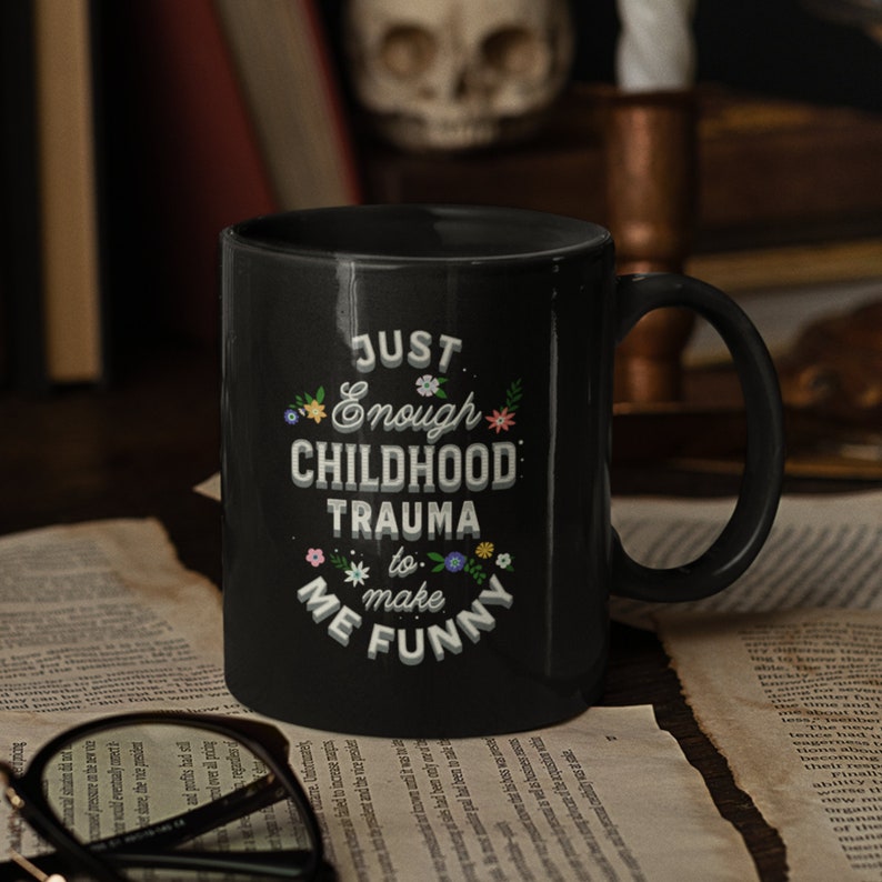 Puede incluir: Taza de caf&eacute; negra con texto blanco que dice "Just Enough Childhood Trauma to Make Me Funny" con peque&ntilde;os detalles florales.