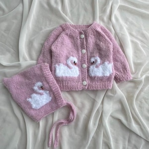 Può includere: Un set di cardigan e cappellino per neonati in maglia rosa chiaro. Il cardigan presenta applicazioni di cigni bianchi e bottoni bianchi. Anche il cappellino ha un'applicazione di cigno. Il set è realizzato in filato morbido e strutturato.