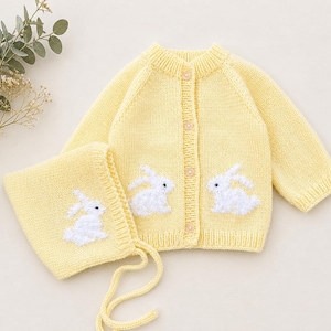 May include: A light yellow knitted baby cardigan and bonnet set. The cardigan features three wooden buttons and two white bunny appliqués. The bonnet has a matching bunny appliqué and ties. The set is made of soft yarn.