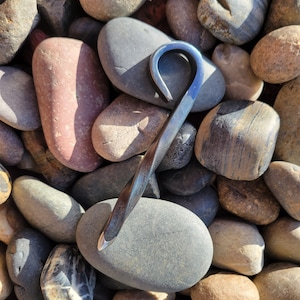 Può includere: Un apribottiglie in metallo forgiato a mano, attorcigliato, con finitura blu e nera. L'apribottiglie ha la forma di un gancio e poggia su un letto di pietre lisce, grigie e marroni.
