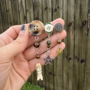 May include: A unique brooch featuring three buttons in shades of brown and green. Dangling from the buttons are beads, a star charm, a leaf, and a small bone wrapped in wire. The brooch is held in a hand against a wooden fence background.