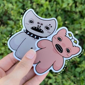 May include: Two cartoon monster stickers. One is gray with a spiked collar and the other is pink with a green plant on its head.