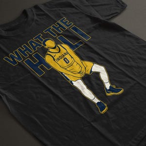 May include: Black t-shirt with a basketball player in a yellow and blue uniform. The text "WHAT THE HELL" is in blue and yellow. The player wears the number 0 and the word "INDIANA" is on the jersey.