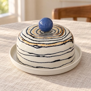 Puede incluir: Una mantequillera de cerámica con tapa en forma de cúpula, base color crema, líneas horizontales en azul marino y dorado metálico, y un asa esférica azul. El recipiente se asienta sobre una base de plato circular a juego color crema.