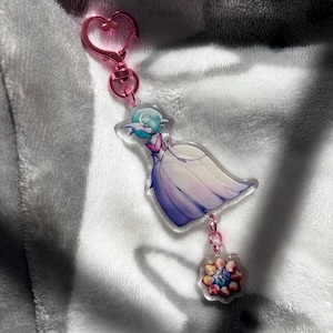 Może przedstawiać: Różowy brelok w kształcie serca z przezroczystym zawieszką przedstawiającą postać z kreskówki w białej sukience i niebieskimi włosami. Zawieszka jest przymocowana do mniejszej, przezroczystej zawieszki z kolorowym wzorem kwiatu.