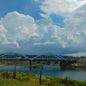 May include: A blue metal bridge spans a river with a cloudy sky overhead. The river is calm and reflects the sky.