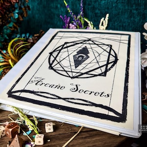 May include: A white binder with a black and white illustration of a geometric design with an eye in the center. The text "Arcane Secrets" is written in cursive below the design.
