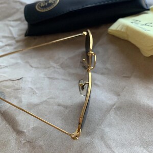 K&ouml;nnte beinhalten: Sonnenbrille mit goldfarbenem Rahmen und dunklen Gl&auml;sern. Die Brille hat ein klassisches Design mit einer d&uuml;nnen, rechteckigen Form. Ein schwarzes Etui und ein gelbes Reinigungstuch befinden sich im Hintergrund.