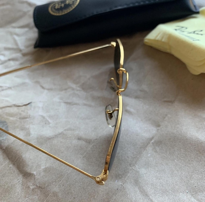 K&ouml;nnte beinhalten: Sonnenbrille mit goldfarbenem Rahmen und dunklen Gl&auml;sern. Die Brille hat ein klassisches Design mit einer d&uuml;nnen, rechteckigen Form. Ein schwarzes Etui und ein gelbes Reinigungstuch befinden sich im Hintergrund.