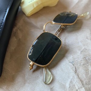 K&ouml;nnte beinhalten: Goldgerahmte Sonnenbrille mit dunklen Gl&auml;sern, mit dem Ray-Ban-Logo auf dem Glas. Die Sonnenbrille ist ge&ouml;ffnet und zeigt die Scharniere und B&uuml;gel. Die Brille liegt auf braunem Papier.