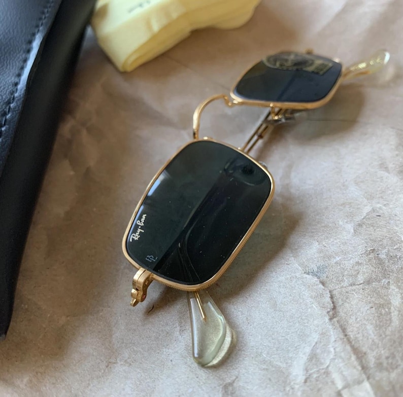 K&ouml;nnte beinhalten: Goldgerahmte Sonnenbrille mit dunklen Gl&auml;sern, mit dem Ray-Ban-Logo auf dem Glas. Die Sonnenbrille ist ge&ouml;ffnet und zeigt die Scharniere und B&uuml;gel. Die Brille liegt auf braunem Papier.