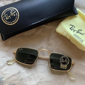 K&ouml;nnte beinhalten: Goldgerahmte Ray-Ban Sonnenbrille mit dunklen Gl&auml;sern, einem schwarzen Etui und einem gelben Reinigungstuch. Die Sonnenbrille hat eine rechteckige Form und das Ray-Ban-Logo ist sichtbar.