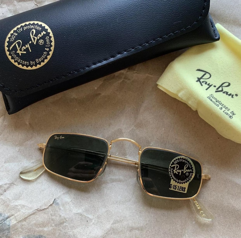 K&ouml;nnte beinhalten: Goldgerahmte Ray-Ban Sonnenbrille mit dunklen Gl&auml;sern, einem schwarzen Etui und einem gelben Reinigungstuch. Die Sonnenbrille hat eine rechteckige Form und das Ray-Ban-Logo ist sichtbar.