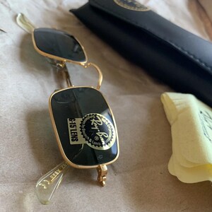 K&ouml;nnte beinhalten: Goldgerahmte Sonnenbrille mit dunkelgr&uuml;nen Gl&auml;sern, einem schwarzen Etui und einem gelben Reinigungstuch. Die Sonnenbrille hat einen Aufkleber mit der Aufschrift "G-15 LENS" und "Ray-Ban". Das Etui ist schwarz und hat ein goldenes Logo.