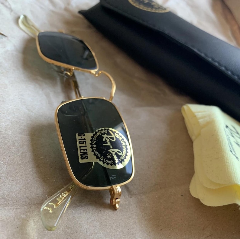 K&ouml;nnte beinhalten: Goldgerahmte Sonnenbrille mit dunkelgr&uuml;nen Gl&auml;sern, einem schwarzen Etui und einem gelben Reinigungstuch. Die Sonnenbrille hat einen Aufkleber mit der Aufschrift "G-15 LENS" und "Ray-Ban". Das Etui ist schwarz und hat ein goldenes Logo.