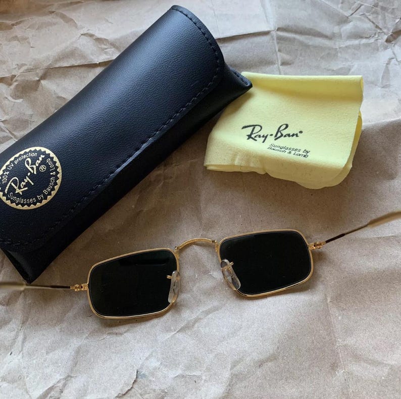 K&ouml;nnte beinhalten: Goldgerahmte Ray-Ban-Sonnenbrille mit rechteckigen schwarzen Gl&auml;sern, einem schwarzen Lederetui und einem gelben Reinigungstuch. Das Etui und das Tuch tragen das Ray-Ban-Logo. Auf einer braunen Papieroberfl&auml;che.