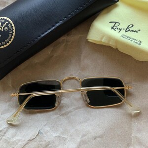 K&ouml;nnte beinhalten: Goldfarbene Ray-Ban Sonnenbrille mit rechteckigen schwarzen Gl&auml;sern. Die Brille wird mit einem schwarzen Etui und einem gelben Reinigungstuch mit Ray-Ban-Logo pr&auml;sentiert. Auf einer hellbraunen Oberfl&auml;che.