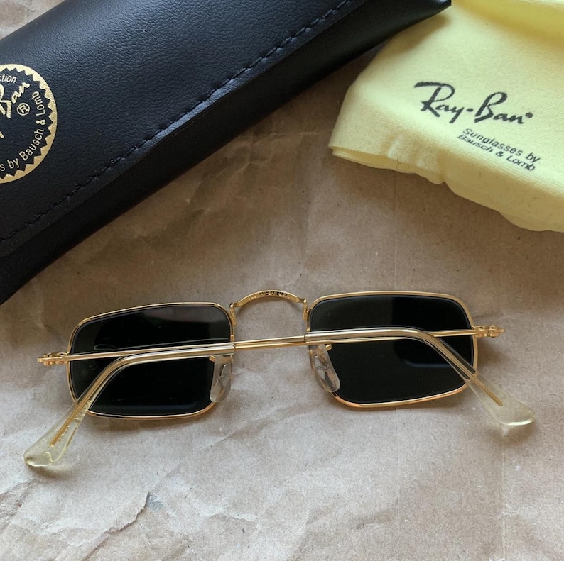 K&ouml;nnte beinhalten: Goldfarbene Ray-Ban Sonnenbrille mit rechteckigen schwarzen Gl&auml;sern. Die Brille wird mit einem schwarzen Etui und einem gelben Reinigungstuch mit Ray-Ban-Logo pr&auml;sentiert. Auf einer hellbraunen Oberfl&auml;che.