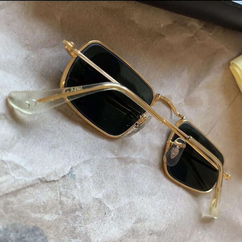 K&ouml;nnte beinhalten: Sonnenbrille mit goldfarbenem Rahmen und rechteckigen schwarzen Gl&auml;sern. Die Sonnenbrille hat ein klassisches Design mit einem d&uuml;nnen Goldrahmen und transparenten B&uuml;gelenden. Die Gl&auml;ser sind dunkel und spenden Schatten. Die Brille ist angewinkelt.