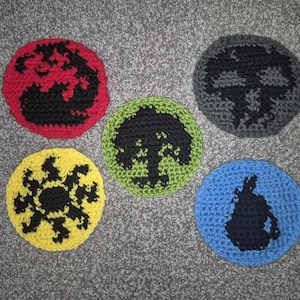 Puede incluir: Cinco círculos de ganchillo en rojo, amarillo, verde, gris y azul. Cada círculo presenta un diseño gráfico negro. Estos artículos hechos a mano son probablemente posavasos o elementos decorativos.