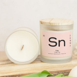 May include: A frosted glass candle jar with a wooden lid. The candle is white and has a gold wick. The label on the jar is pink and says "Sn TIN 118.710 Hand Poured | Blended Soy Wax | Made in USA".