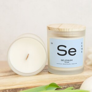 May include: A frosted glass candle jar with a wooden lid. The candle is white and has a wooden wick. The label on the jar is blue and says "Se Selenium 78.96 Hand Poured | Blended Soy Wax | Made in USA".