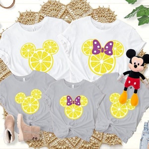 May include: Four t-shirts featuring a lemon-shaped Mickey Mouse head design. Two shirts are white with a purple bow on the Minnie Mouse head, and two shirts are gray with a purple bow on the Minnie Mouse head.
