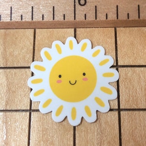 May include: A white and yellow sticker with a smiling sun face. The sun has a yellow face with pink cheeks and a closed-mouth smile. The sun is surrounded by white petals with yellow centers.