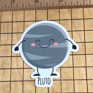 May include: A cartoon illustration of Pluto, a dwarf planet, with a smiling face and small arms and legs. The planet is grey with a white background and the word "PLUTO" is written below.