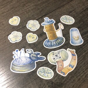May include: A set of 10 blue and yellow illustrated sewing themed stickers. The stickers include a spool of thread with a bird on top, a thimble with a bird on the side, a pincushion with pins, a measuring tape, and various buttons.
