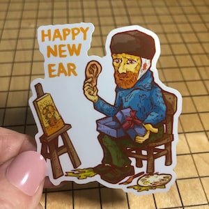 Könnte beinhalten: Ein Aufkleber mit einer Karikatur von Vincent van Gogh, der ein abgetrenntes Ohr und eine Geschenkbox hält. Der Text "Happy New Ear" ist in orangefarbenen Buchstaben oben auf dem Aufkleber geschrieben.