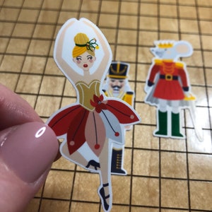May include: A ballerina sticker with a red and white tutu, a gold bodice, and a yellow bun. The ballerina is on her toes with her arms raised above her head.