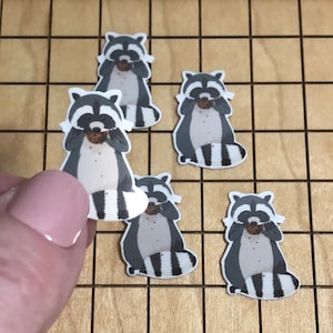 May include: A collection of raccoon stickers, each featuring a gray and white raccoon covering its eyes with a cookie. The stickers have a white border and are arranged on a light brown grid.