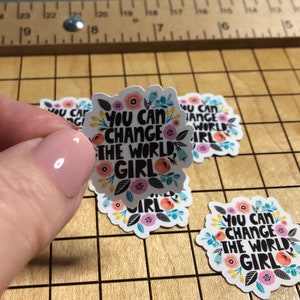 May include: Three round stickers with a floral design and the text "You can change the world, girl".
