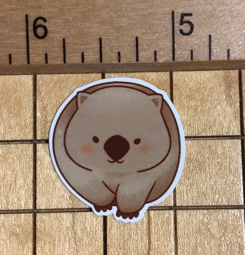 Puede incluir: Una pegatina de wombat de dibujos animados marr&oacute;n y blanco con una expresi&oacute;n linda. La pegatina es redonda y tiene un contorno blanco.