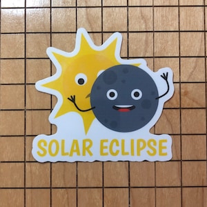 May include: A white sticker featuring a yellow sun and a grey moon. The sun and moon have faces, with the moon partially covering the sun. The text "SOLAR ECLIPSE" is positioned below the sun and moon.