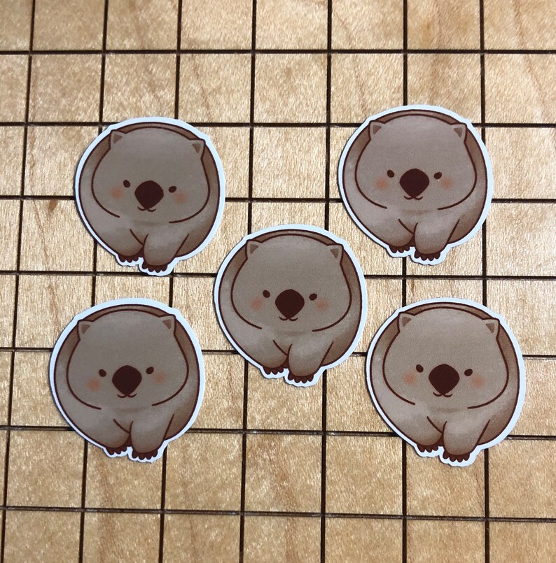 Puede incluir: Cinco pegatinas de wombat de dibujos animados marrones y blancas sobre un fondo de madera. Los wombats est&aacute;n todos mirando hacia adelante con la boca cerrada y los ojos cerrados. Todos llevan un abrigo de piel marr&oacute;n y blanco.