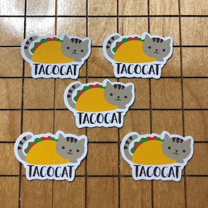 May include: Five gray cat stickers with the word "Tacocat" printed on each. Each cat is wearing a yellow taco with green lettuce and red tomato.