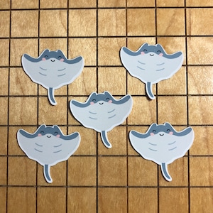 May include: Five gray and blue stingray stickers with smiling faces on a wooden background.