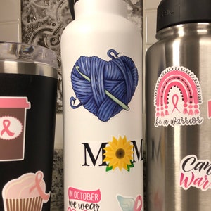 May include: White stainless steel water bottle with various stickers. Stickers include a heart-shaped ball of yarn with a knitting needle, a sunflower, the words "MOM" in black, a pink ribbon with wings, and the text "In October we wear pink".