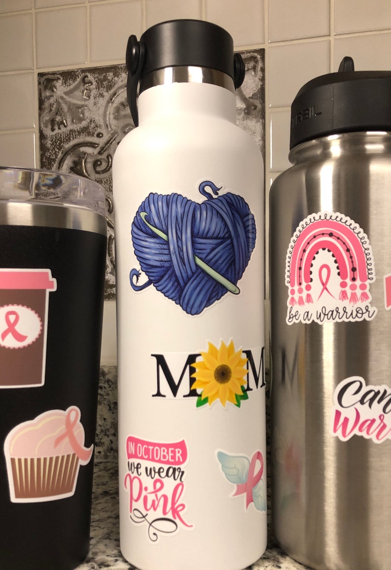 May include: White stainless steel water bottle with various stickers. Stickers include a heart-shaped ball of yarn with a knitting needle, a sunflower, the words "MOM" in black, a pink ribbon with wings, and the text "In October we wear pink".