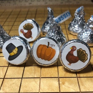 May include: Thanksgiving-themed Hershey's Kisses candies. Each candy is wrapped in silver foil and topped with a circular sticker. Stickers feature a turkey, a pilgrim hat, and a pumpkin. The word "KISSES" is visible on a few of the candy wrappers.