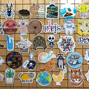 May include: A collection of colorful stickers featuring various designs, including animals, plants, and text. Some of the stickers have a humorous or inspirational message. The stickers are arranged on a wooden surface.