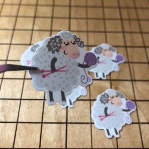May include: Three white sheep stickers with black eyes and pink noses. The sheep are holding purple yarn and knitting needles. The sheep are on a brown and white checkered background.