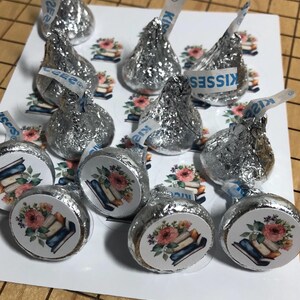 May include: Silver foil-wrapped chocolate candies with white paper strips that read "Kisses." The candies are arranged on a white surface with floral and book-themed stickers. The stickers feature a stack of books and pink flowers.