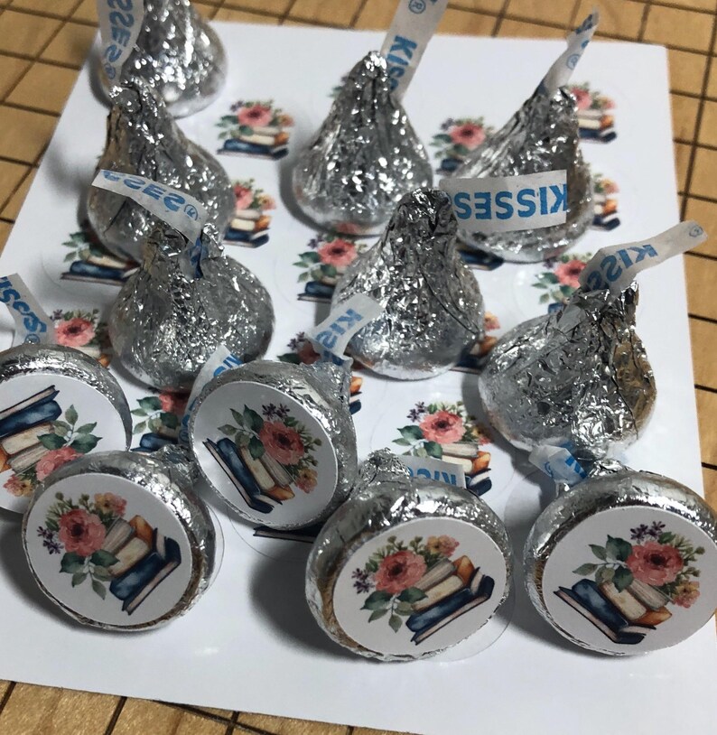 May include: Silver foil-wrapped chocolate candies with white paper strips that read "Kisses." The candies are arranged on a white surface with floral and book-themed stickers. The stickers feature a stack of books and pink flowers.