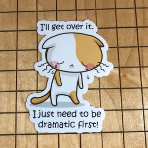May include: A white and orange cartoon cat with a sad expression and teary eyes. The cat is standing on all fours and has a tail. The text "I'll get over it. I just need to be dramatic first!" is written below the cat.