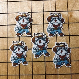 May include: Five cartoon raccoon stickers with the word "Howdy" on each. The raccoons are wearing cowboy hats, bandanas, and jeans. They are all smiling and have a happy expression on their faces.